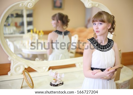 Beautiful girl in white stands near mirror and holds frame with photo in light room.