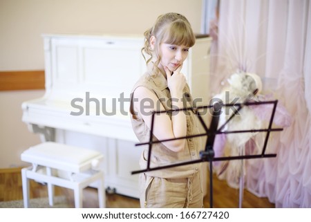 Beautiful girl stands near music stand in room with classic interior and piano.
