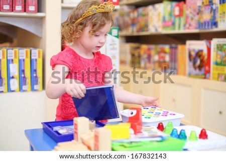Little pretty girl in crown of princess plays with toys in store.