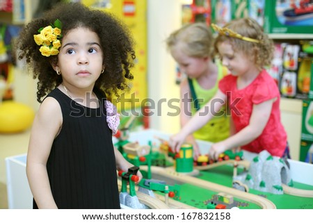 Little girl looks up and two children play with toy railroad in store. Focus on left girl.