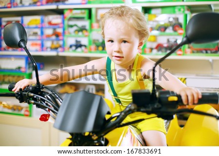 Little cute boy in yellow sits on toy motorcycle in store with toys.