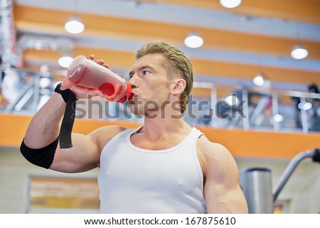 Young athlete man drinks beverage from bottle during  break between exercises in gym hall