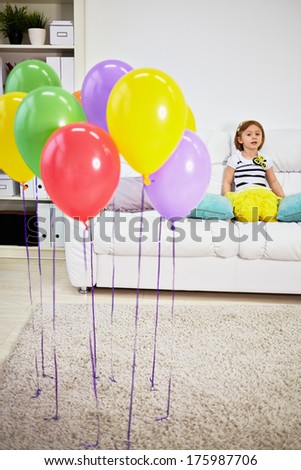 Little girl sits on big sofa in room with light carpet on floor and birthday air balloons