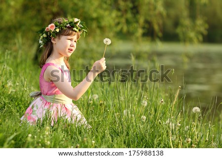 Little pretty girl in pink sits and looks at dandelion on green grass in summer park. Focus on hand and dandelion.