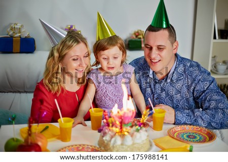 Family of three sits at birthday table, daughter prepares to snuff out candle on cake and parents look at her