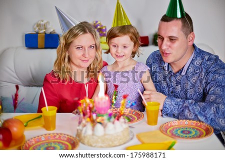 Mother, father and little daughter sit together on soft sofa at birthday table and look at burning candle on cake