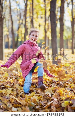 Little girl runs along autumn park kicking up fallen leaves with her boots and jumps