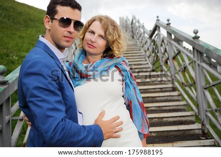 Pregnant woman in white stands on stairs and stylish man touches her belly outdoor. Focus on woman.