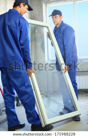 Two workers in blue work clothes carry the window to install it in the frame
