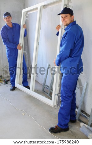 Two men in work clothes holding new window frame in a room under renovation