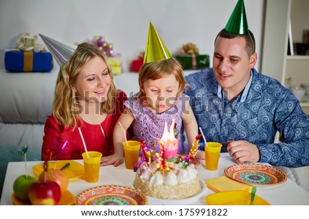 Family of three sits at birthday table, daughter snuffes out candle on cake and parents look at her