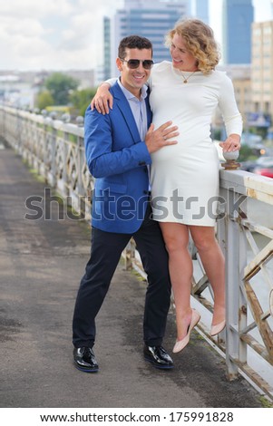 Pregnant woman in white stands on railings and stylish man touches her belly outdoor in city.