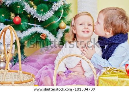 Two children sit in room under christmas tree on furry rug, boy whispers something in girl ear