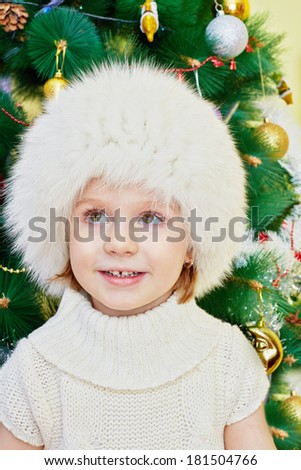 Portrait of little girl dressed in knitted dress and fur hat with christmas tree at background