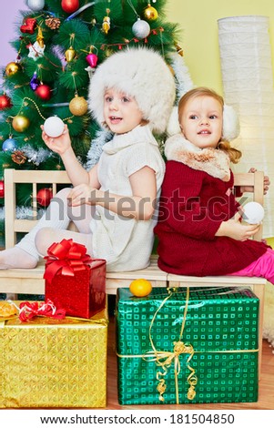 Two little girls sit on wooden bench back to back with decorated artificial new year tree at background in room
