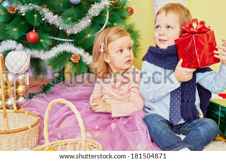 Little girl and boy sit on furry rug under christmas tree, boy holds red gift box, girl look at him