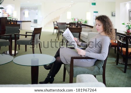 Pretty woman in dress sits at table in hall and reads newspaper