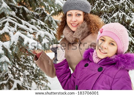 Mother and daughter stand under snow-covered firtree on winter day