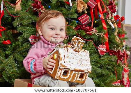 Little girl sits on big cardboard gift box with contented look under Christmas tree, holding gingerbread house in her hands