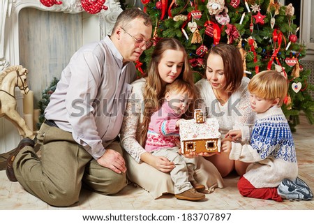 Family of five sits under Christmas tree and looks over gingerbread house in hands of mother, younger children touch it