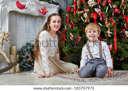 Teenage girl and little boy sit on floor under Christmas tree, boy sits among burning tealights which stand in shape of heart