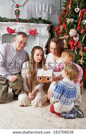 Family of five sits under Christmas tree and looks over gingerbread house in hands of mother