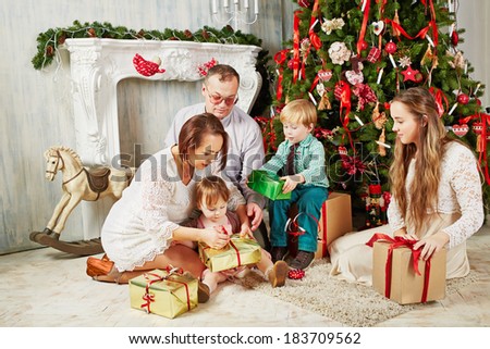 Parents and children sit on rug near Christmas tree, untying ribbons on gift boxes