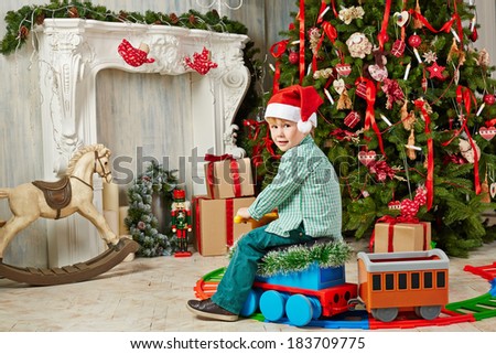 Little boy in Santa cap sits on toy plastic steam engine at room decorated to Christmas celebration