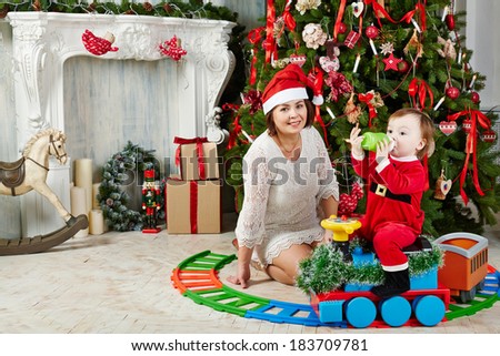 Mother in santa cap sits on floor under decorated Christmas tree near little girl dressed in Santa suit which sits on toy plastic steam engine and drink from bottle