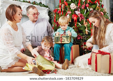Family of five sits under Christmas tree, parents look at their children, wich untie gift boxes