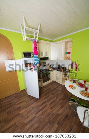 Little girl upside down holds a fridge in the kitchen with table and dishes