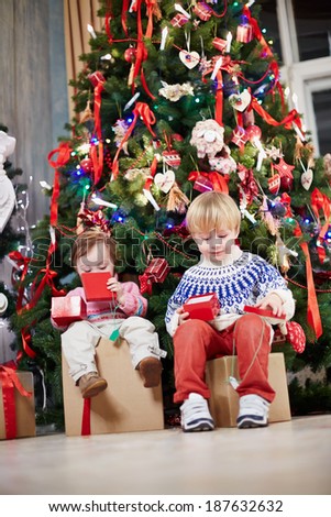 Little boy and little girl sit on big cardboard gift boxes under Christmas tree, holding open gift boxes of smaller size