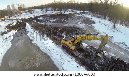 Excavators on dirt snow-covered ground reduced pond near the forest, view from unmanned quadrocopter.