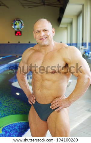 Happy bodybuilder in swimming trunks stands near indoor pool of gym hall
