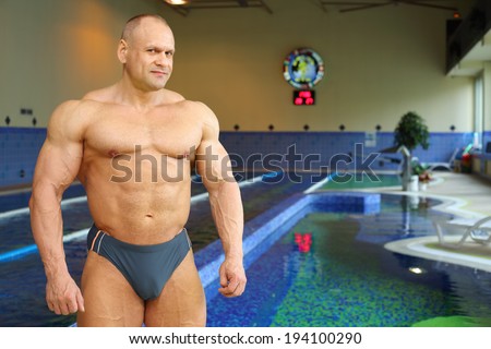 Bodybuilder in swimming trunks stands near indoor pool of gym hall