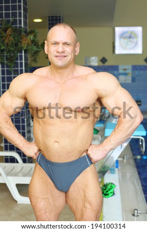 Happy sunburnt bodybuilder stands near indoor pool in big modern gym hall