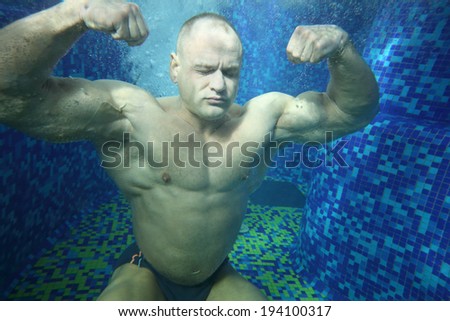 Bodybuilder in swimming trunks sits on bottom of pool underwater among many bubbles