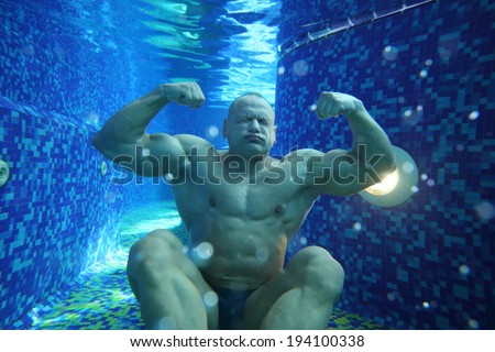 Bodybuilder in swimming trunks sits on bottom of pool underwater with pure water