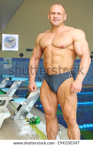 Happy sunburnt bodybuilder stands near indoor pool before swimming in gym hall