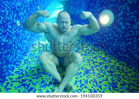 Bodybuilder in swimming trunks sits on bottom of pool underwater