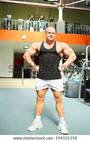 Bodybuilder in black jersey and white jogging shoes stands in gym hall and smiles