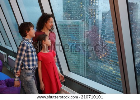 Mother with two children looking at evening city through a large window