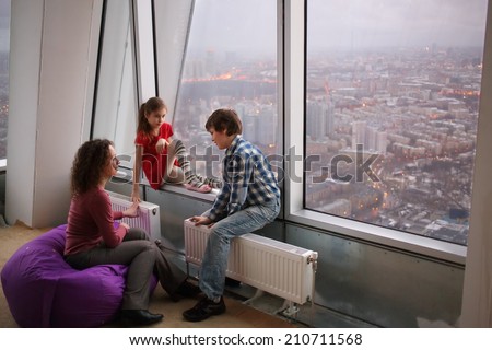 Woman and two children sitting next to a large window with a view of the evening city