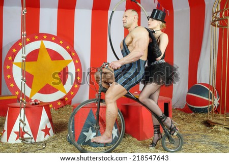 Two circus performers musclemen and girl ride on retro bike in striped tent