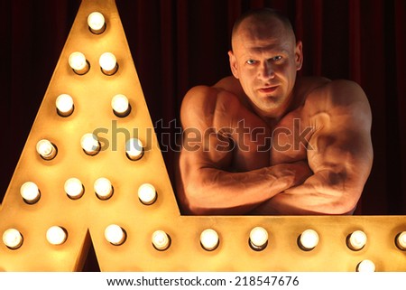 Strong bodybuilder with naked torso leaning on big star with lights
