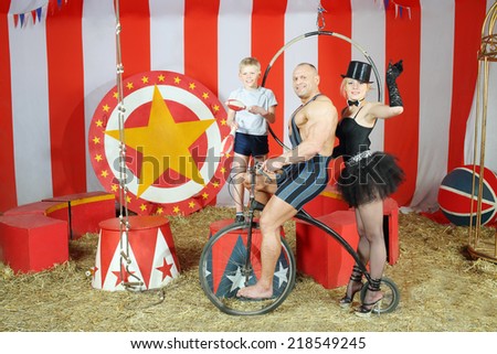 Three funny circus strong man, woman in cylinder and boy on retro bike with balloons in striped tent