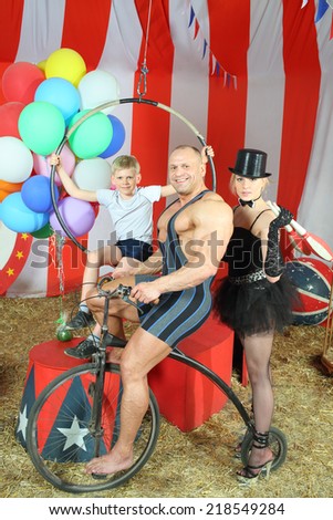 Three happy circus strong man, woman in cylinder and boy on retro bike with balloons in striped tent
