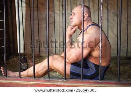 Sad strongman sits with his head by hand in the cage for animals