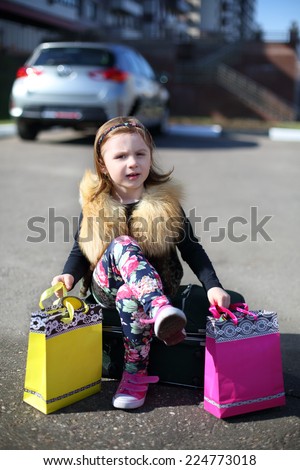 Little girl sitting on suitcase with her legs crossed with colored gift bags in middle of road