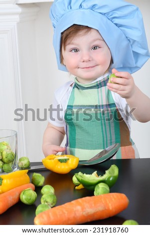 Beautiful little girl in kitchen apron and cap play at table with vegetables